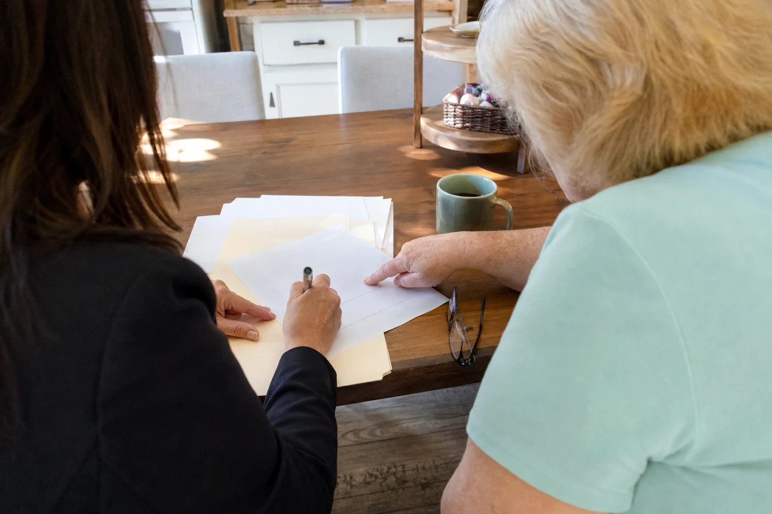 Agent in a dark jacket writing on papers at a wooden kitchen table beside an older woman, with a green coffee mug and reading glasses on the table