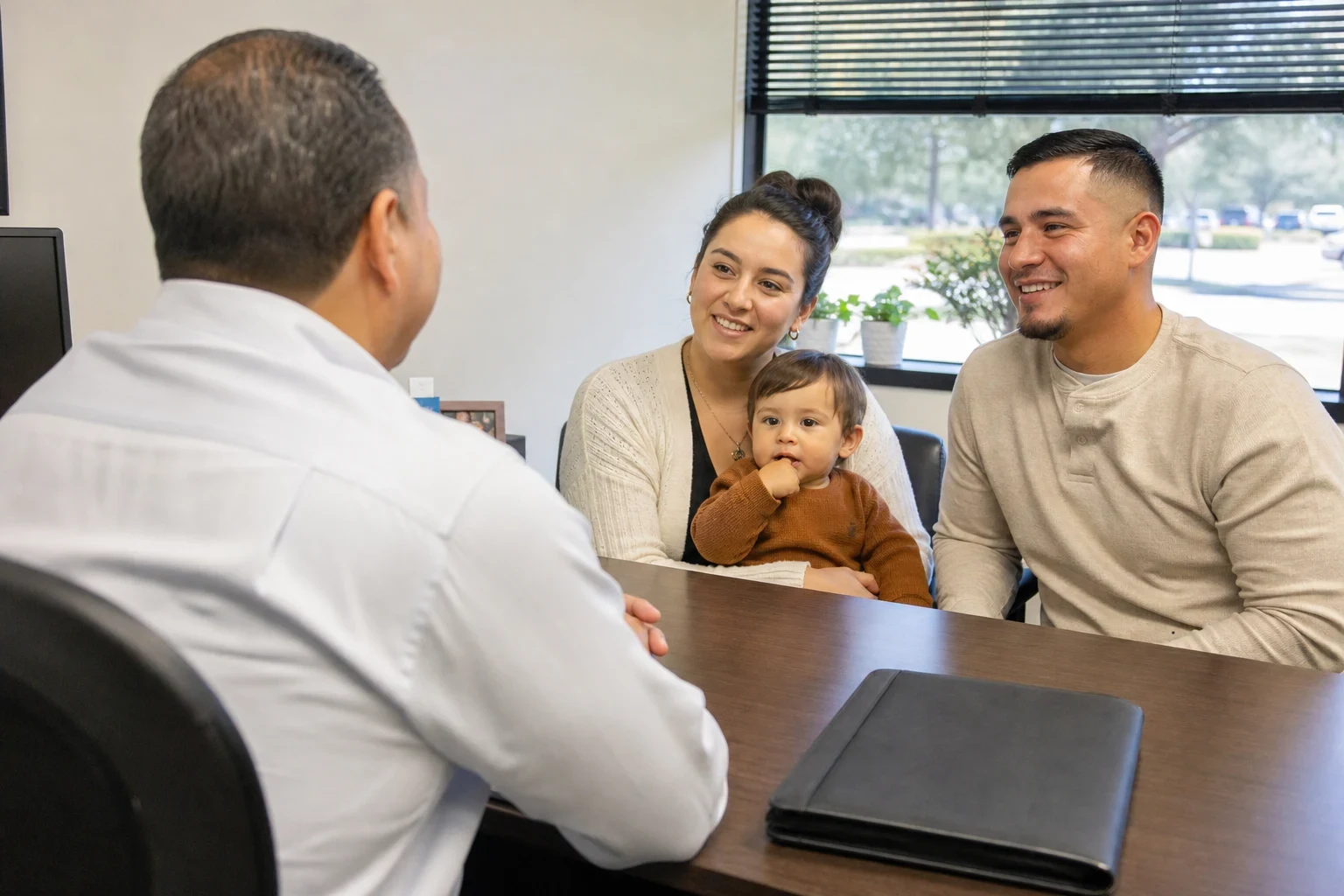 Insurance agent seen from behind talking with a smiling couple and their toddler at a wooden office desk with a closed black folder, window behind them