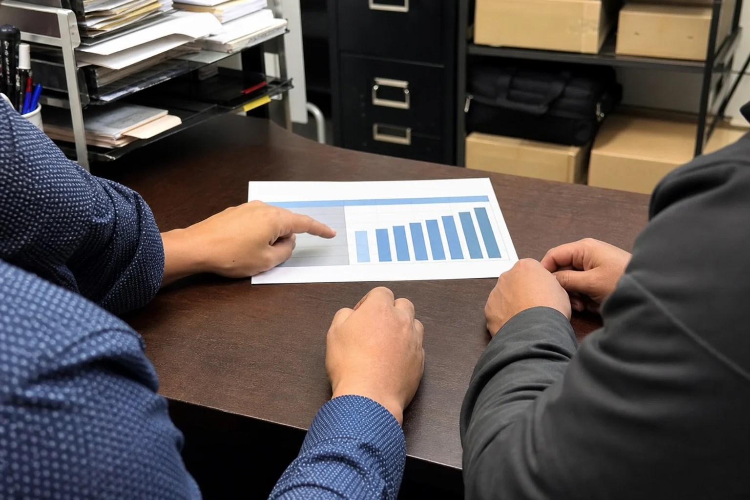 Two people seated at a dark wood desk in a back office, one pointing to a printed bar chart, with paper trays, a black filing cabinet and boxes on shelves behind